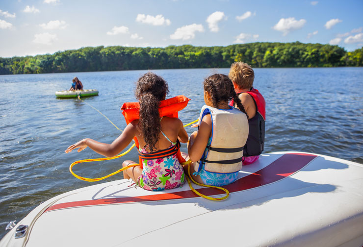 three kids sitting on a ski boat on a lake hooked to a tube